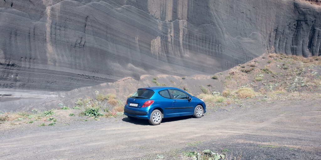 A blue three-door hatchback photographed from the rear three-quarter view, with a black volcanic formation visible in the background.