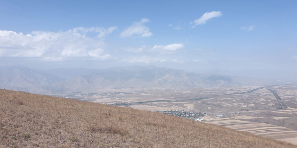 A mountainous landscape visible from on top of a mountain through a bit of fog, with a cloudy sky visible at the top of the image.