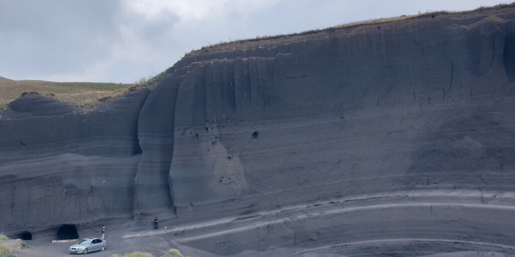 A black wall made of volcanic rock with layers of lighter-colored rock running across. A silver car is visible in the distance right next to the wall, while the top parts of the image show a cloudy sky.