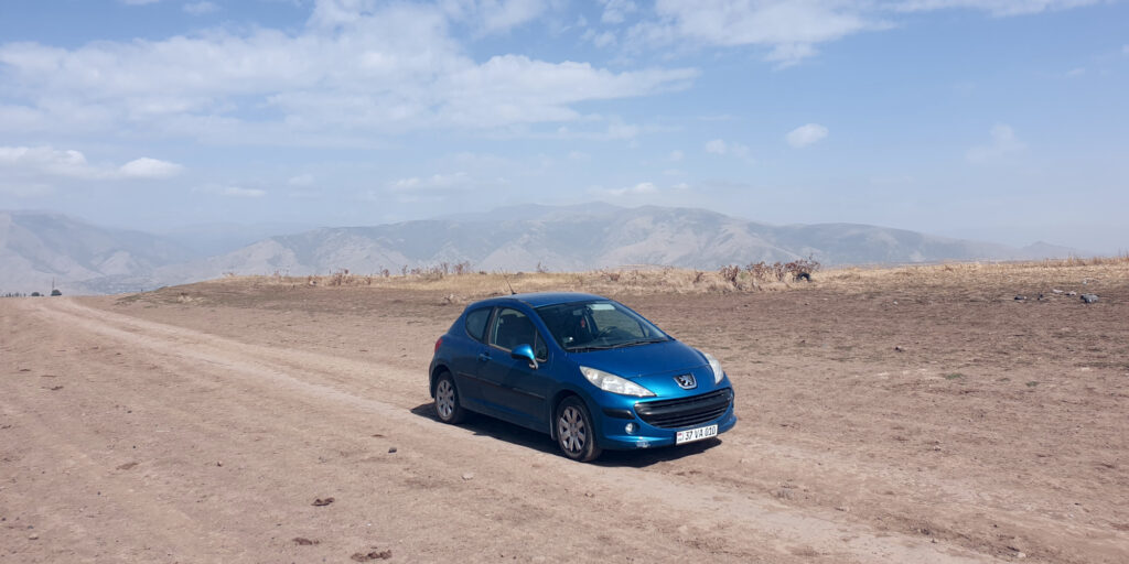 A wide-angle photo of a blue hatchback car from the right-front three-quarter view in the middle of a wide dirt trail. In the background, mountains are visible through the fog, while the upper part of the image shows a cloudy sky.