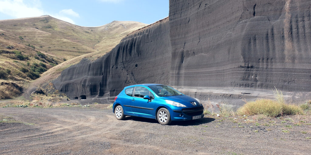 A wide shot of a blue hatchback car from the right-front three-quarter view. The car is on a gravel/dirt surface, with bits of grass visible to the right. The background is a black volcanic wall across the majority of the image, with a mountain peak visible on the left of the image.