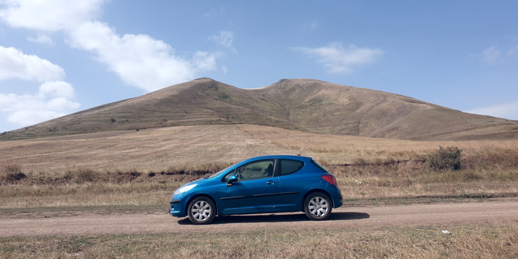 A wide-angle photo of a blue hatchback car photographed from its left side. The car is on a dirt trail, with some small shrubs visible in the closer parts of the background. The upper part of the image shows the peak of a mountain, with a cloudy sky visible above it.