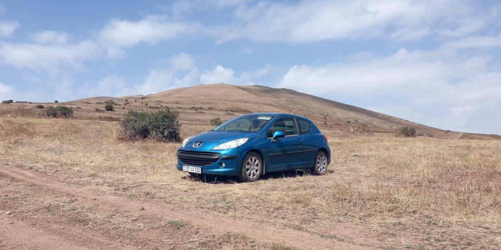 A wide-angle photo of a blue car, a Peugeot 207, from its front-left three-quarter view. The car is in a field, right next to a dirt path. There’s a rosehip bush visible to the left of the car in the background, with the rest of the background showing a mountain. The upper parts of the image show a cloudy sky.