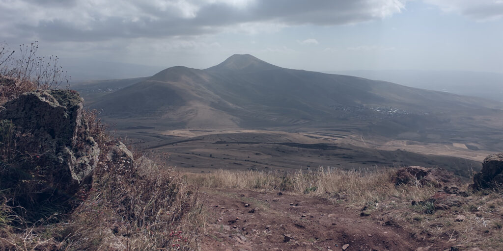 A photo of a mountain shot from a dirt path at the peak of another mountain of the same size. The mountain, which is visible in the mid-to-upper parts of the image, is covered in blotchy shadows from low-flying clouds, which are visible in the upper parts of the image. The foreground of the image shows a dirt trail with lots of small rocks. To the left of the trail, there is a larger rock visible.