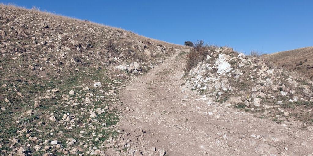 A photo of a dirt path going up the side of a steep hill. The path is covered in small rocks all over, with larger rocks being visible on either side of the path, all across the hillside. A clear blue sky is visible in the top parts of the image.